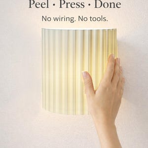 May include: A cream-colored, cylindrical wall lamp with vertical ridges is mounted on a light pink wall. The lamp is illuminated from within, and a hand is touching the lamp. The text "Peel • Press • Done" and "No wiring. No tools." is above the lamp.