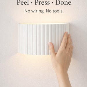 May include: A white, cylindrical wall sconce with vertical ridges is being installed on a white wall. The text "Peel • Press • Done" and "No wiring. No tools." is above the sconce.