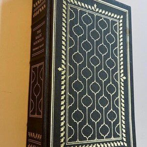May include: A hardcover book titled "The Magic Mountain" by Thomas Mann. The book has a dark cover with gold decorative accents and a repeating geometric pattern. The spine and edges are also dark with gold detailing.