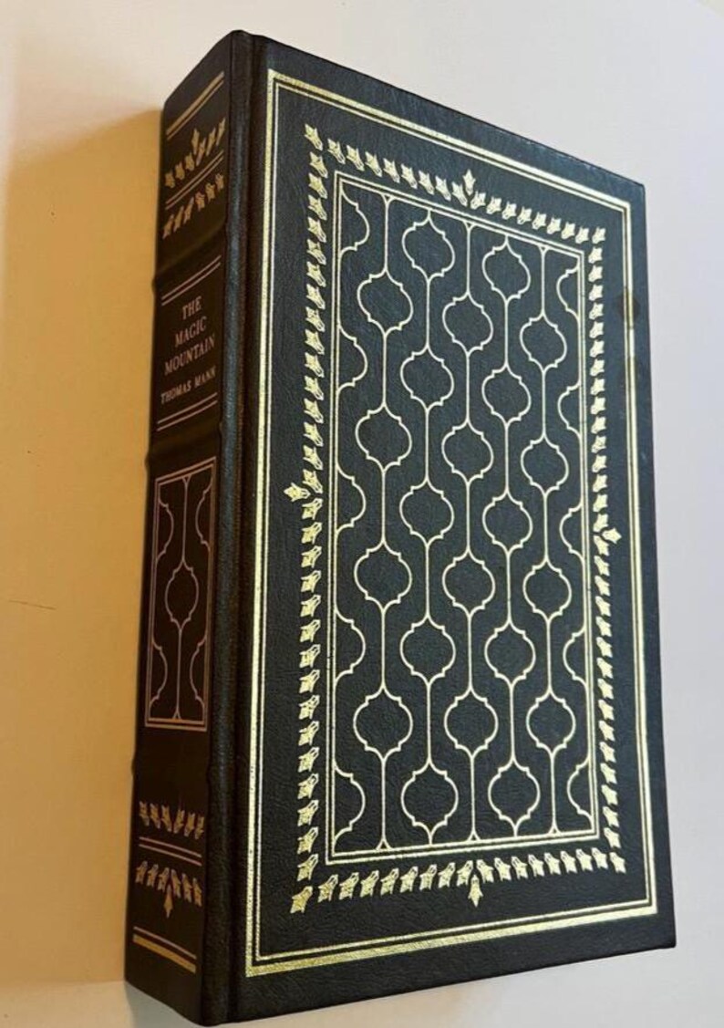 May include: A hardcover book titled "The Magic Mountain" by Thomas Mann. The book has a dark cover with gold decorative accents and a repeating geometric pattern. The spine and edges are also dark with gold detailing.