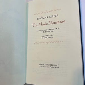 May include: Open book with the title "The Magic Mountain" by Thomas Mann. The text is in a serif font and is printed in red and black ink. The book is from The Franklin Library, Franklin Center, Pennsylvania.