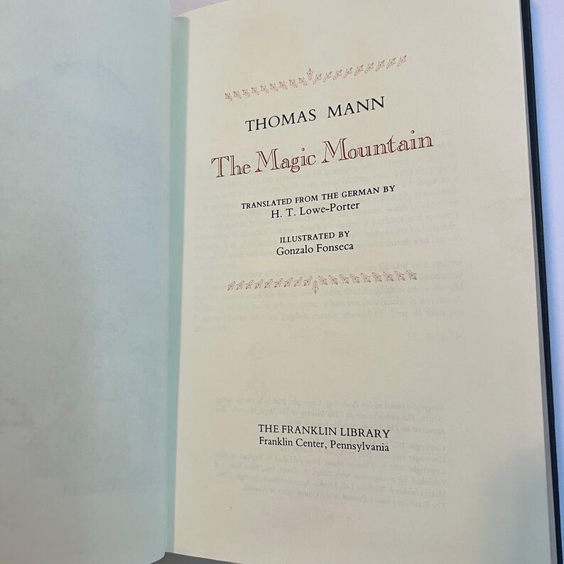 May include: Open book with the title "The Magic Mountain" by Thomas Mann. The text is in a serif font and is printed in red and black ink. The book is from The Franklin Library, Franklin Center, Pennsylvania.