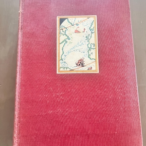 May include: A vintage book with a textured, red cover. A rectangular illustration is centered on the cover, depicting a map with a ship and a creature. The book's spine is visible, and the edges show signs of wear.
