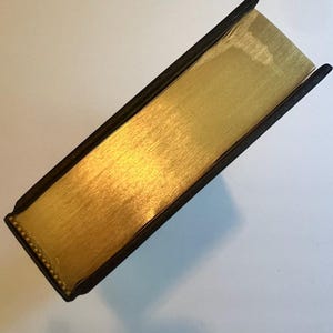 May include: A close-up view of a book with gold-gilded edges. The book's pages are a bright gold color, and the edges are smooth and reflective. The cover is black with a textured appearance.