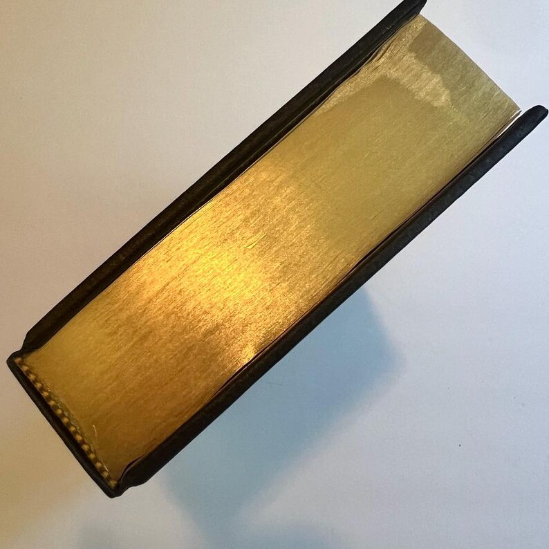 May include: A close-up view of a book with gold-gilded edges. The book's pages are a bright gold color, and the edges are smooth and reflective. The cover is black with a textured appearance.