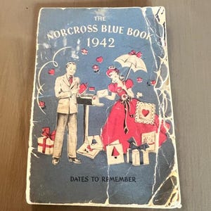 Puede incluir: Libro azul Norcross vintage de 1942 con una cubierta azul. La portada presenta una pareja, regalos y corazones. El título del libro y el año están en texto blanco. La frase "Dates to Remember" está en la parte inferior.
