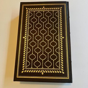 May include: A hardcover book with a dark brown cover and gold accents. The cover features a gold geometric pattern and a gold border with a leaf design. The book's spine is also dark brown.