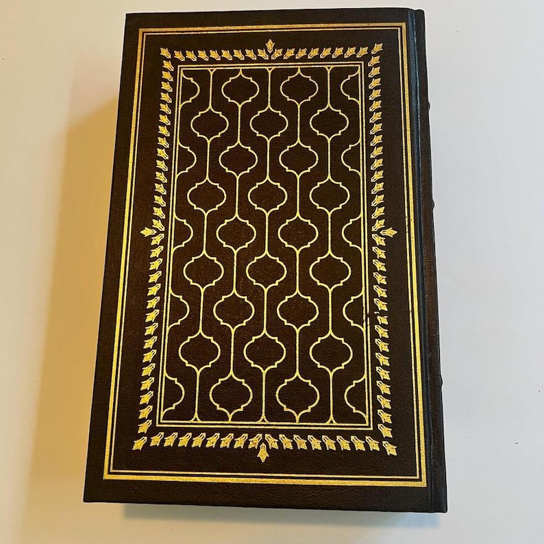 May include: A hardcover book with a dark brown cover and gold accents. The cover features a gold geometric pattern and a gold border with a leaf design. The book's spine is also dark brown.