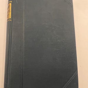 Könnte beinhalten: Ein Vintage-Hardcover-Buch mit dunkelblauem Einband. Der Buchrücken hat goldene Schrift und einen goldenen Rand. Der Einband hat ein strukturiertes Aussehen und ein diagonales Liniendesign an den Ecken.