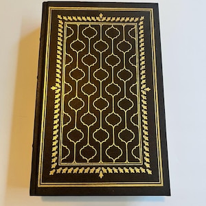 May include: A hardcover book with a dark brown cover and gold decorative accents. The cover features a gold geometric pattern and a gold border with leaf-like designs. The book's spine is also dark brown.