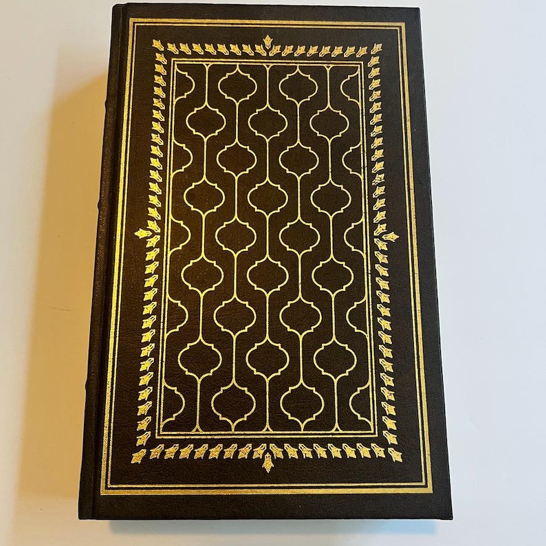 May include: A hardcover book with a dark brown cover and gold decorative accents. The cover features a gold geometric pattern and a gold border with leaf-like designs. The book's spine is also dark brown.