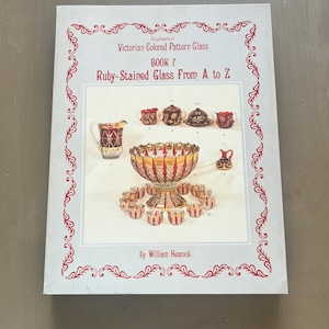May include: A book titled "Victorian Colored Pattern Glass Book 7: Ruby-Stained Glass From A to Z" features illustrations of ruby-stained glassware. The cover has a light blue background with a red decorative border and text.