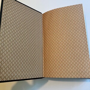May include: Open book with a black cover and tan pages. The pages have a repeating pattern of small white crosses. The book is open to reveal two pages with the same pattern, one in a darker tan and the other in a lighter tan.