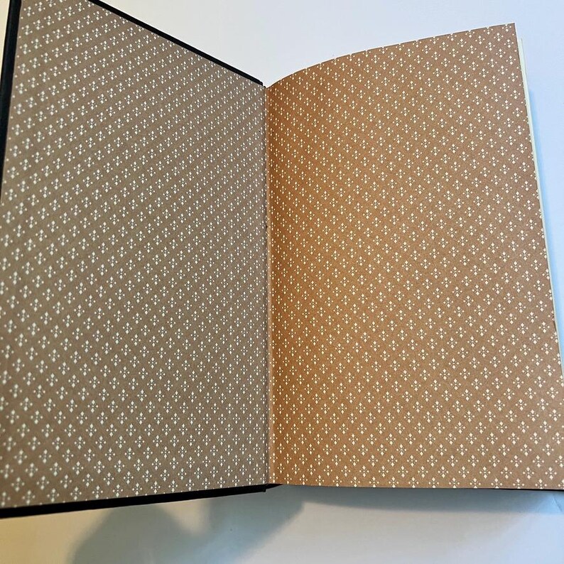 May include: Open book with a black cover and tan pages. The pages have a repeating pattern of small white crosses. The book is open to reveal two pages with the same pattern, one in a darker tan and the other in a lighter tan.