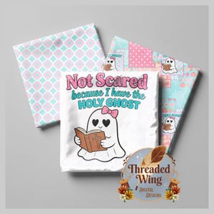 May include: A white t-shirt featuring a cartoon ghost with heart-shaped eyes and a pink bow, reading a brown book with a cross. The text reads Not Scared because I have the HOLY GHOST. Two folded fabric swatches with blue and pink patterns are behind.