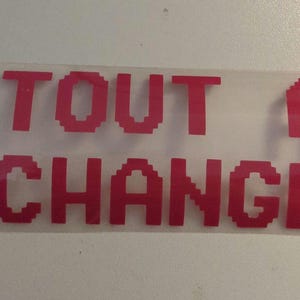 May include: A clear, transparent transfer sheet with the words "TOUT A CHANGÉ" in pixelated, bright pink letters. The text is arranged in two lines, with the first line reading "TOUT A" and the second line reading "CHANGÉ".