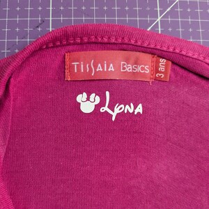 Peut inclure: Gros plan sur un t-shirt magenta. Le t-shirt porte une étiquette rouge avec l'inscription "Ti(s)AIA Basics" et "3 ans". En dessous de l'étiquette se trouve un graphisme blanc et le nom "LpNA".