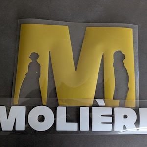 Op de afbeelding: Een gouden vinyl sticker met de letter "M" en twee silhouetten van mensen. Onder de letter staat het woord "MOLIERE" in wit vinyl.