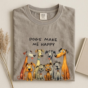 Peut inclure: T-shirt gris avec l'inscription noire « DOGS MAKE ME HAPPY ». En dessous, une illustration colorée de huit chiens de dessin animé. Le t-shirt a un col rond et des manches courtes.
