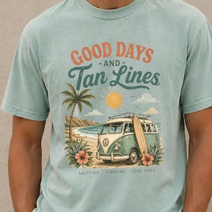 May include: A light blue t-shirt featuring a graphic print of a vintage van on a beach with a surfboard, a palm tree, and a sun. The text reads GOOD DAYS AND Tan Lines, with SALTY AIR - SUNSHINE - GOOD VIBES printed in smaller letters below the van.