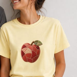 Op de afbeelding: Een lichtgeel t-shirt met korte mouwen en een centrale appelafbeelding in een patchwork-ontwerp. De appel is samengesteld uit verschillende tinten rode en bruine stoftexturen met een groen blad met patroon aan de steel.