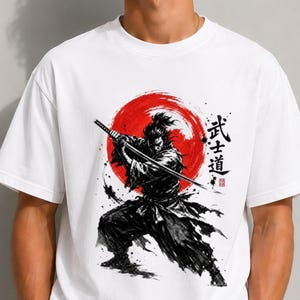Op de afbeelding: Een wit T-shirt met korte mouwen en een zwarte inktillustratie van een samoerai die een katana vasthoudt. Achter de figuur bevindt zich een grote rode cirkel. Rechts van de samoerai staan zwarte Japanse karakters en een klein rood stempel.