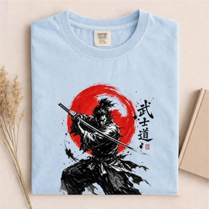 Op de afbeelding: Een lichtblauw t-shirt met een grafische print van een samoerai die een katana vasthoudt voor een grote rode zon. Het ontwerp bevat zwarte inktachtige penseelstreken en Japanse kanji-tekens die Bushido lezen, met een Comfort Colors-label.