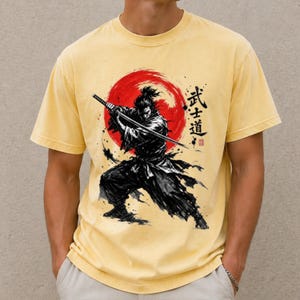 Op de afbeelding: Een lichtgeel T-shirt met korte mouwen en een zwarte inkt-stijl illustratie van een samoerai met een katana. Het ontwerp bevat een grote rode cirkelvormige zon op de achtergrond en verticale Japanse kanji-tekens rechts van de figuur.