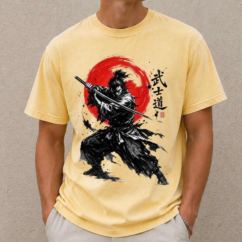 Op de afbeelding: Een lichtgeel T-shirt met korte mouwen en een zwarte inkt-stijl illustratie van een samoerai met een katana. Het ontwerp bevat een grote rode cirkelvormige zon op de achtergrond en verticale Japanse kanji-tekens rechts van de figuur.