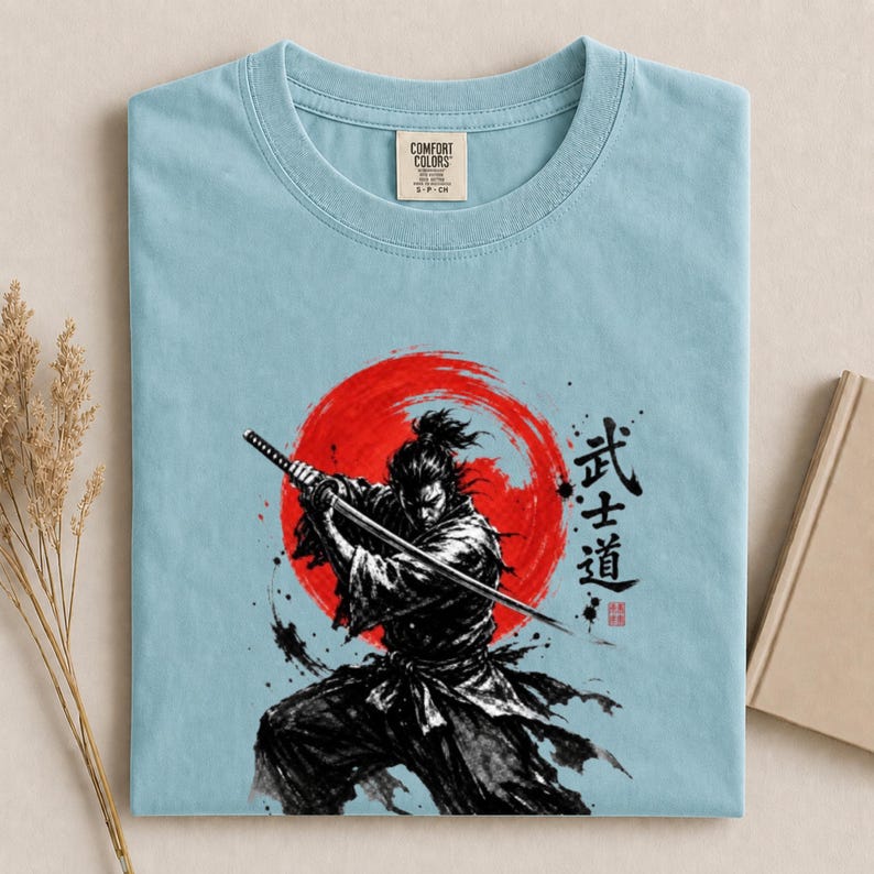 Op de afbeelding: Een lichtblauw t-shirt met een afbeelding van een samoerai die een katana vasthoudt voor een grote rode zon. Het ontwerp bevat zwarte penseelstreken in inktstijl en Japanse kanji-tekens. Het shirt heeft een Comfort Colors neklabel.