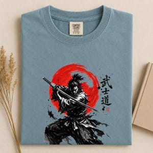 Op de afbeelding: Een stoffig blauw t-shirt met een grafische print van een samoerai die een katana vasthoudt voor een grote rode zon. Het ontwerp bevat zwarte penseelstreken in inktstijl en Japanse kanji-tekens rechts van de figuur. Het t-shirt heeft een Comfort Colors-merklabel bij de binnenhals.