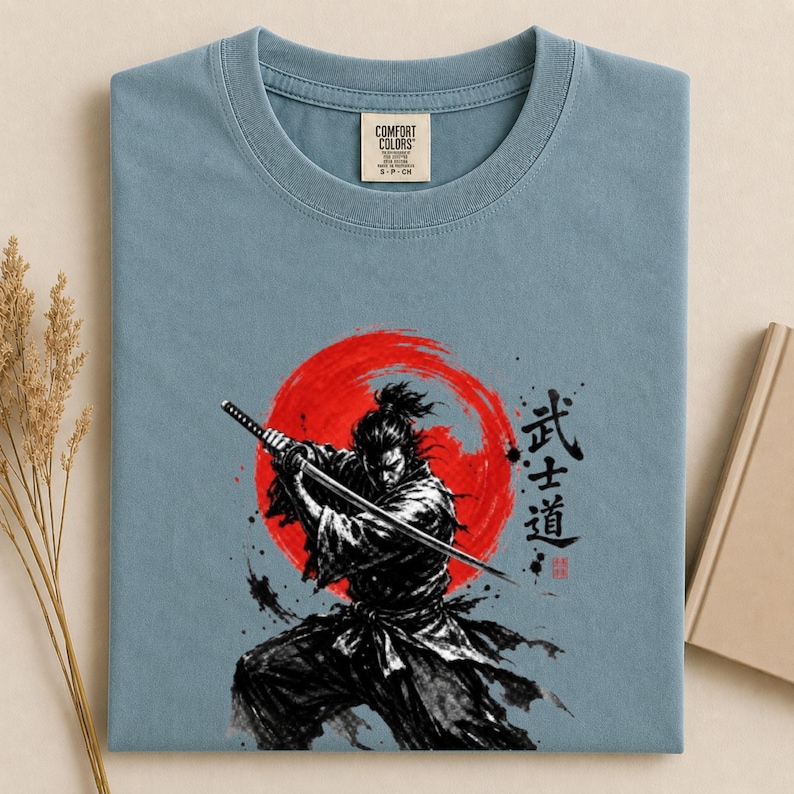 Op de afbeelding: Een stoffig blauw t-shirt met een grafische print van een samoerai die een katana vasthoudt voor een grote rode zon. Het ontwerp bevat zwarte penseelstreken in inktstijl en Japanse kanji-tekens rechts van de figuur. Het t-shirt heeft een Comfort Colors-merklabel bij de binnenhals.