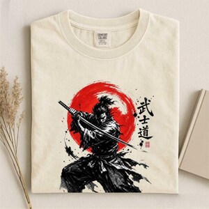 Op de afbeelding: Een cr&egrave;mekleurig t-shirt met een afbeelding van een samoerai die een katana vasthoudt voor een grote rode cirkel. Het ontwerp bevat zwarte penseelstreken in inktstijl en Japanse kanji-tekens rechts van de figuur, met een Comfort Colors label.