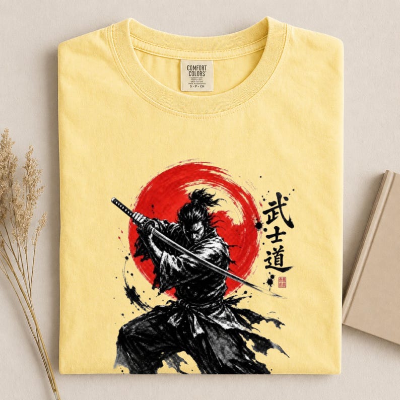 Op de afbeelding: Een lichtgeel t-shirt met een grafische print van een samoerai die een katana in een dynamische houding vasthoudt. Achter het figuur staat een grote rode cirkel. Rechts van de samoerai staan zwarte Japanse kanji en een kleine rode stempel.