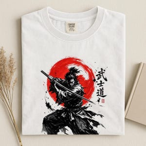 Op de afbeelding: Een wit t-shirt met een zwarte inktillustratie van een samoerai die een katana vasthoudt. Achter het figuur bevindt zich een groot rood cirkelvormig zonmotief. Rechts staan zwarte Japanse kanji verticaal gedrukt met een klein rood zegel.