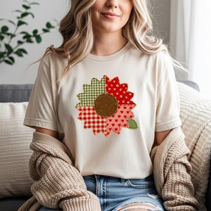 May include: A cream-coloured short-sleeve t-shirt featuring a large sunflower graphic on the chest. The sunflower petals are divided into four distinct sections with different patterns: green gingham, red floral, red gingham, and pink heart print.