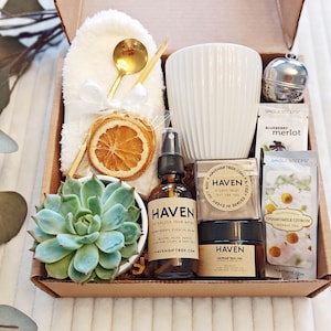 May include: A gift box filled with self-care items, including a white mug, a small succulent plant, a bottle of essential oil, a jar of face cream, a tea infuser, and tea bags. The box also includes a white hand towel, a gold spoon, and a dried orange slice.