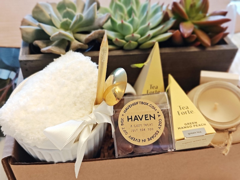 K&ouml;nnte beinhalten: Eine Geschenkbox gef&uuml;llt mit einem wei&szlig;en Handtuch, einer kleinen Tasse, einem goldenen L&ouml;ffel, einer Schachtel Pralinen, einer Kerze und zwei Teebeuteln. Die Box ist mit einem braunen Etikett verziert, auf dem "Haven: A Cozy Treat Just For You" steht.