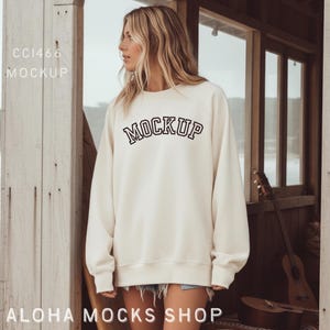 May include: Cream-colored sweatshirt with the word "MOCKUP" in black, outlined letters. The sweatshirt is long-sleeved and worn by a person. The text "CC1466 MOCKUP" is in the upper left corner, and "ALOHA MOCKS SHOP" is at the bottom.