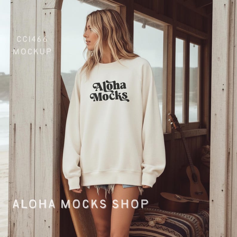 May include: Cream-colored sweatshirt with the text "Aloha Mocks" in a black, retro font. The sweatshirt is long-sleeved and has a relaxed fit. The image is taken outdoors, with a wooden structure in the background.
