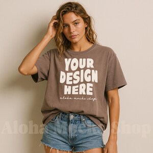 May include: A brown t-shirt with the text "YOUR DESIGN HERE" in large white block letters. The text "aloha mocks shop" is below the main text. The model is wearing blue denim shorts.