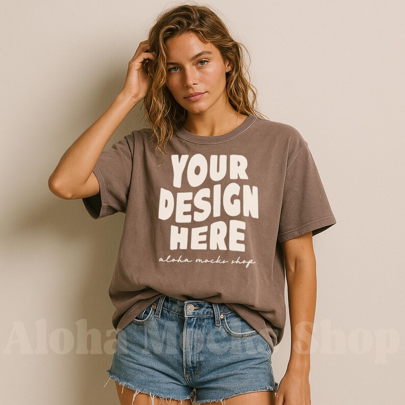 May include: A brown t-shirt with the text "YOUR DESIGN HERE" in large white block letters. The text "aloha mocks shop" is below the main text. The model is wearing blue denim shorts.