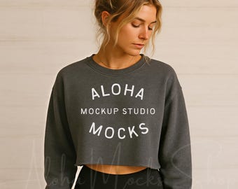 May include: A dark gray cropped sweatshirt with the text "ALOHA MOCKUP STUDIO MOCKS" in white. The sweatshirt has a crew neck and a raw hem. The model is wearing black leggings. The background is a light wood panel.
