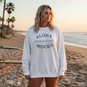 May include: White sweatshirt with the text "Aloha Mockup Studio Mocks" in gray. The sweatshirt is worn by a person on a beach with palm trees and the ocean in the background. The image is taken during sunset.