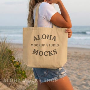May include: A beige tote bag with the words "ALOHA MOCKUP STUDIO MOCKS" printed in dark gray. The bag has long straps and is held by a person. The words "ALOHA MOCKS SHOP" are printed at the bottom.