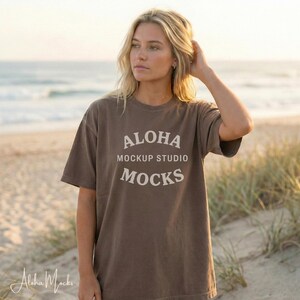 May include: A brown t-shirt with the text "ALOHA MOCKUP STUDIO MOCKS" in white. The t-shirt is worn by a person on a beach. The person is looking off to the side. The background includes the ocean and sand.