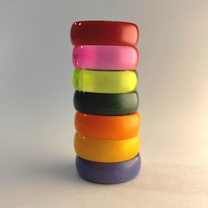 Op de afbeelding: Een stapel van zeven kleurrijke armbanden in rood, roze, groen, zwart, oranje, geel en paars. Elke armband is effen van kleur en heeft een gladde, afgeronde vorm. De armbanden zijn verticaal gestapeld tegen een witte achtergrond.