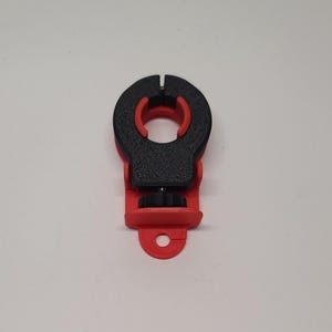 May include: A black and red plastic clamp with a screw adjustment. The clamp has a circular opening and a small hole at the bottom.