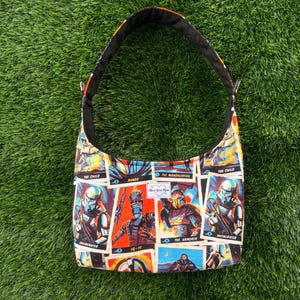 Pu&ograve; includere: Una borsa a tracolla con cinturino nero e stampa colorata con personaggi di una serie di fantascienza. Il design della borsa include immagini di personaggi come The Mandalorian e The Child, con interno nero.