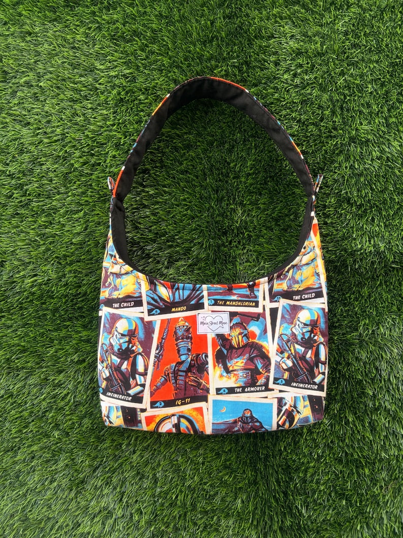 Pu&ograve; includere: Una borsa a tracolla con cinturino nero e stampa colorata con personaggi di una serie di fantascienza. Il design della borsa include immagini di personaggi come The Mandalorian e The Child, con interno nero.